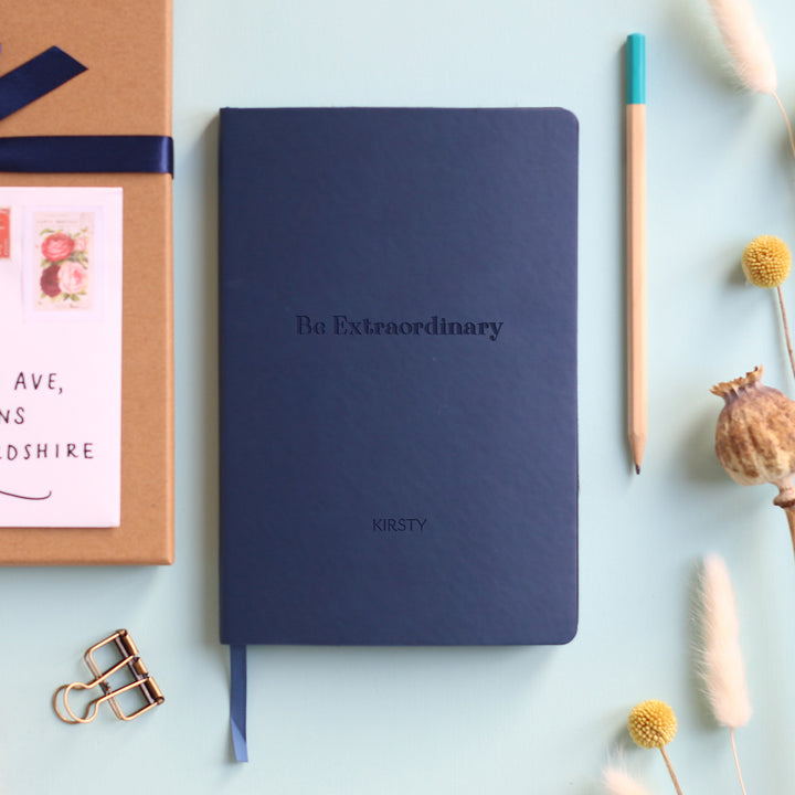 A top down image of a navy blue vegan leather notebook resting on a pale aqua table. The front of the notebook features the words Be Extraordinary. It is personalised with the name Kirsty. Surrounding the book are various stationary items and dried flower heads.