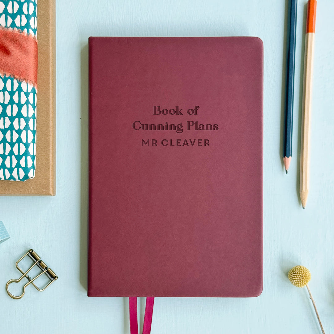 A flat lay image of a burgundy vegan leather notebook resting on a pale aqua table. The front of the notebook is engraved with the phrase “Book of Cunning Plans” and is personalised with the name Mr Cleaver. Surrounding the book are various stationary items.