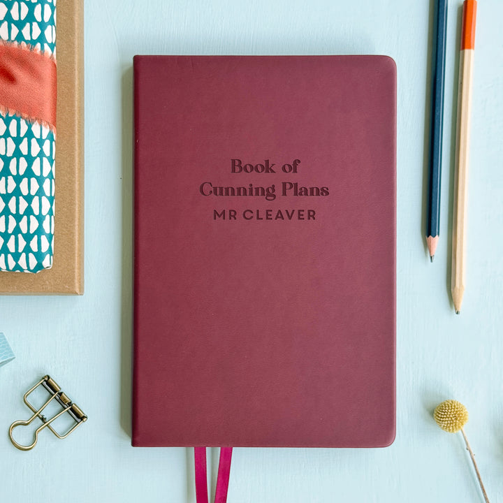 A flat lay image of a burgundy vegan leather notebook resting on a pale aqua table. The front of the notebook is engraved with the phrase “Book of Cunning Plans” and is personalised with the name Mr Cleaver. Surrounding the book are various stationary items.