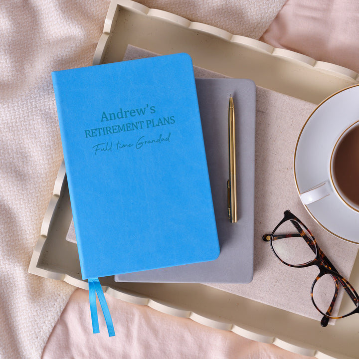 A top down image of a blue notebook on top of a on a grey vegan leather notebook resting on a tray with a pen, a pair of glasses and a cup and saucer containing tea to the right. The front of the notebook is engraved with Andrew's Retirement Plans, followed by a line of personalised text.
