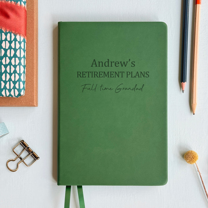 A top down image of an dark green vegan leather notebook resting on a pale aqua table. The front of the notebook is engraved with  Andrew's Retirement Plans followed by a line of personalised text. Surrounding the book are various stationary items and dried flowers.