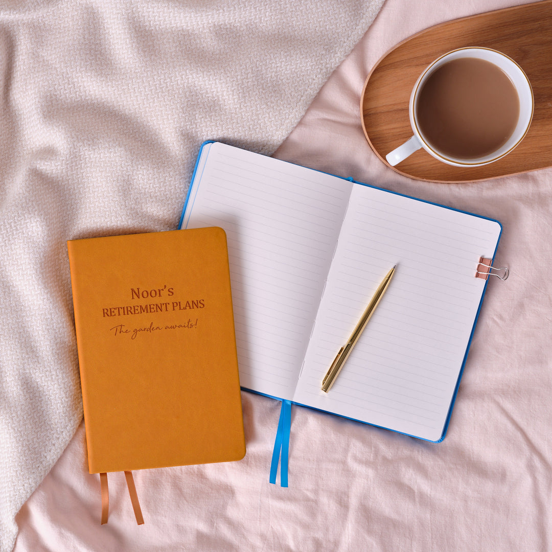 A top down image of a mustard yellow notebook resting on an open blue vegan leather notebook with a pen and a cup and saucer containing tea to the right. The front of the Noor's Retirement Plans, followed by a line of personalised text.