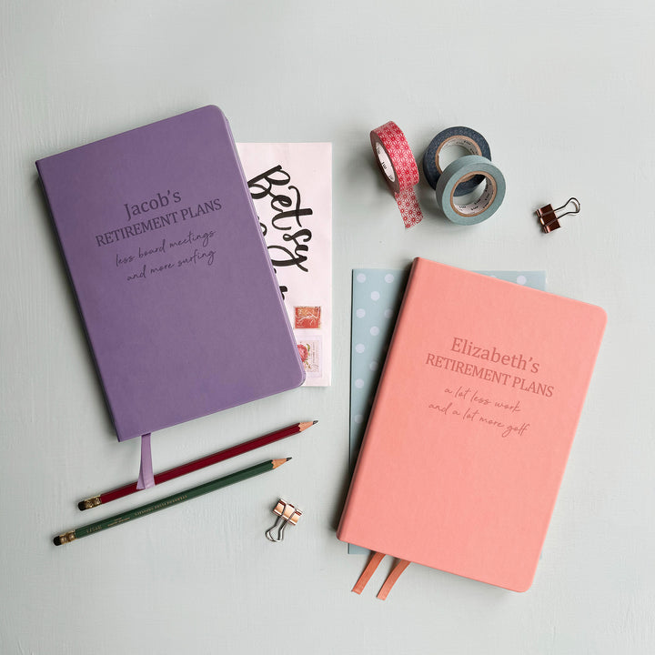 A top down image of two vegan leather notebooks, one lilac and one pink. They are resting on a pale aqua table. The lilac notebook is engraved with the words Jacob's Retirement and the pink book is engraved with the words Elizabeth's Retirement. Both also have an additional line of personalised text. Surrounding the book are various stationary items.