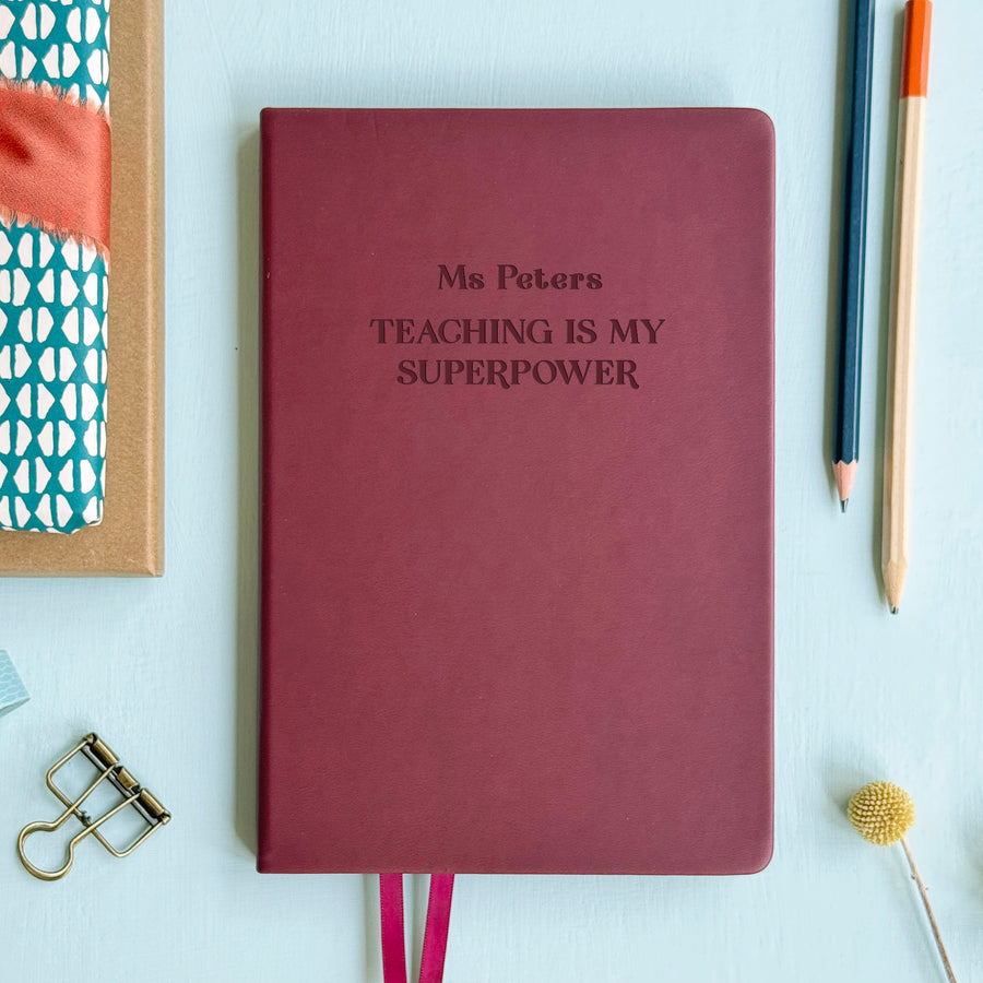 A flat lay image of a burgundy vegan leather notebook resting on a pale aqua table. The front of the notebook is engraved with the phrase “Teaching is my Superpower” and is personalised with the name Ms Peters. Surrounding the book are various stationery items.