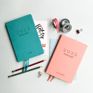 A top down image of two vegan leather notebooks, one turquoise and one pale pink. They are resting on a pale aqua table. The front of each notebook is engraved with 2025 Journal. Personalised with the name Craig on the turquoise notebook and Alexandria is engraved on the pink notebook. Surrounding the book are various stationary items.
