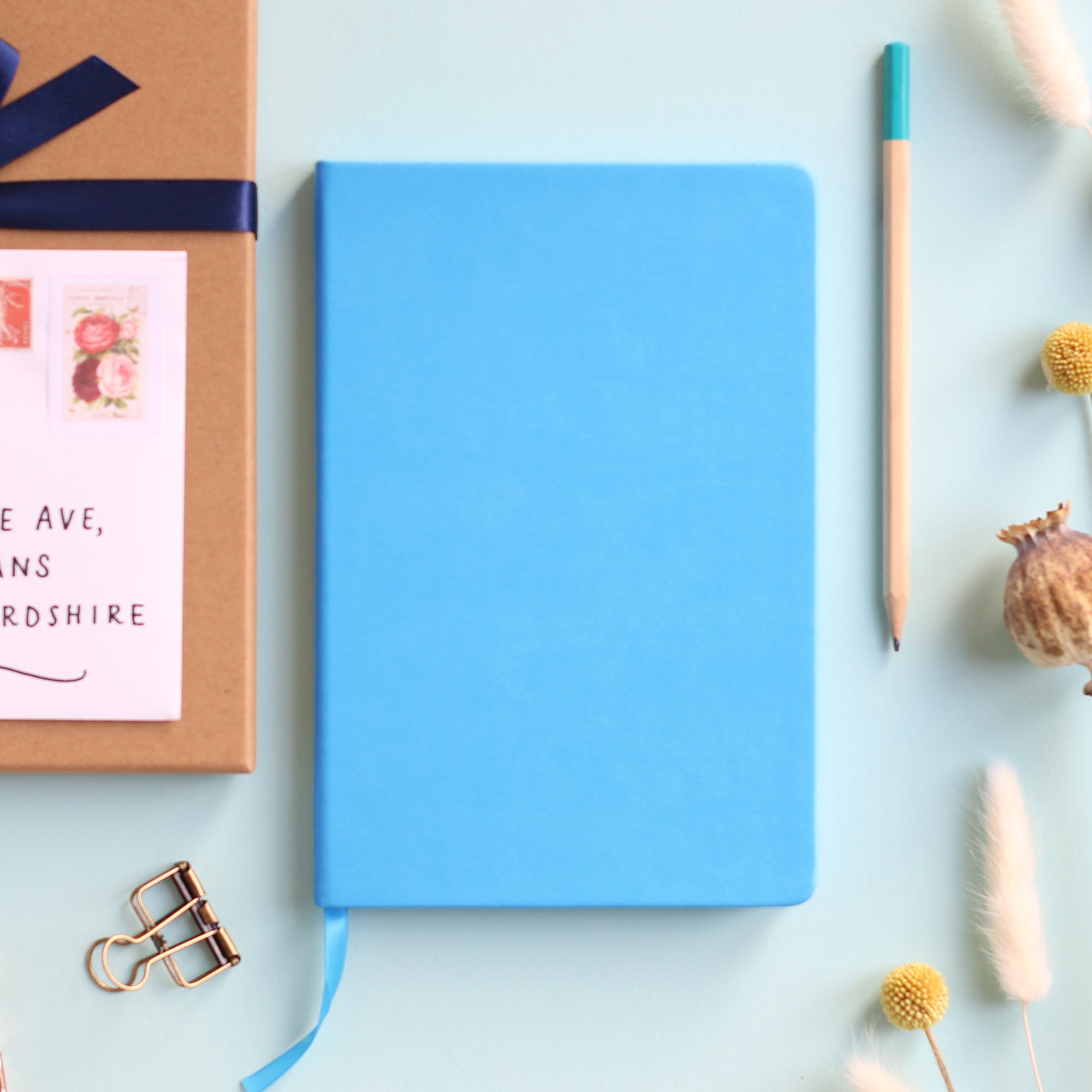 A top down image of a plain simple blue vegan leather notebook. It is resting on a pale aqua table. Surrounding the book are various stationary items and dried flowers.