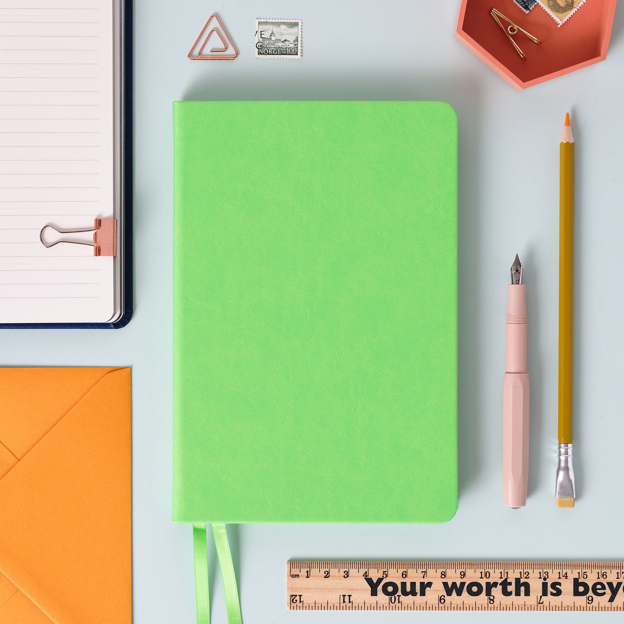 A top down image of a plain simple green vegan leather notebook. It is resting on a pale aqua table. Surrounding the book are various stationary items.