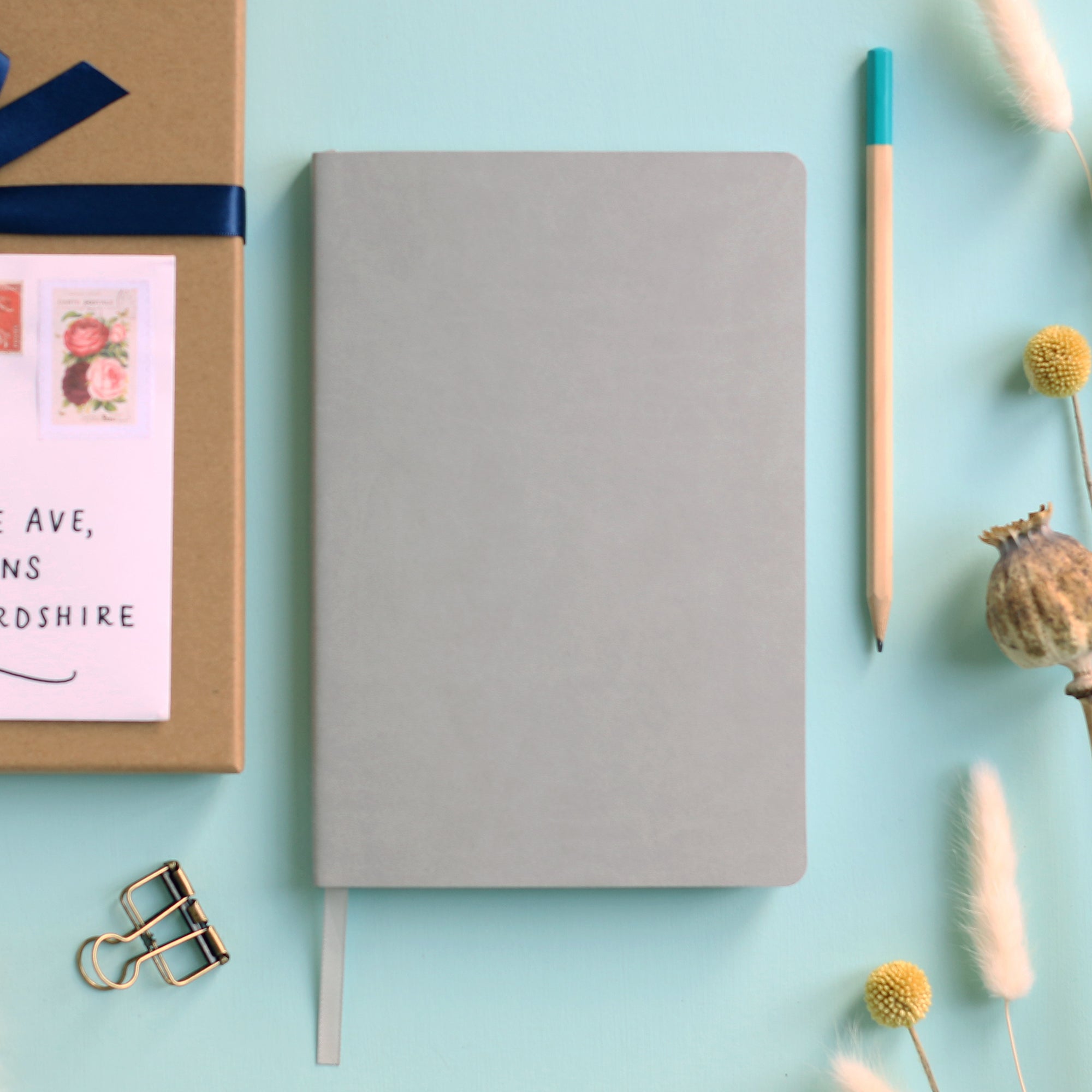 A top down image of a plain simple grey vegan leather notebook. It is resting on a pale aqua table. Surrounding the book are various stationary items and dried flowers.