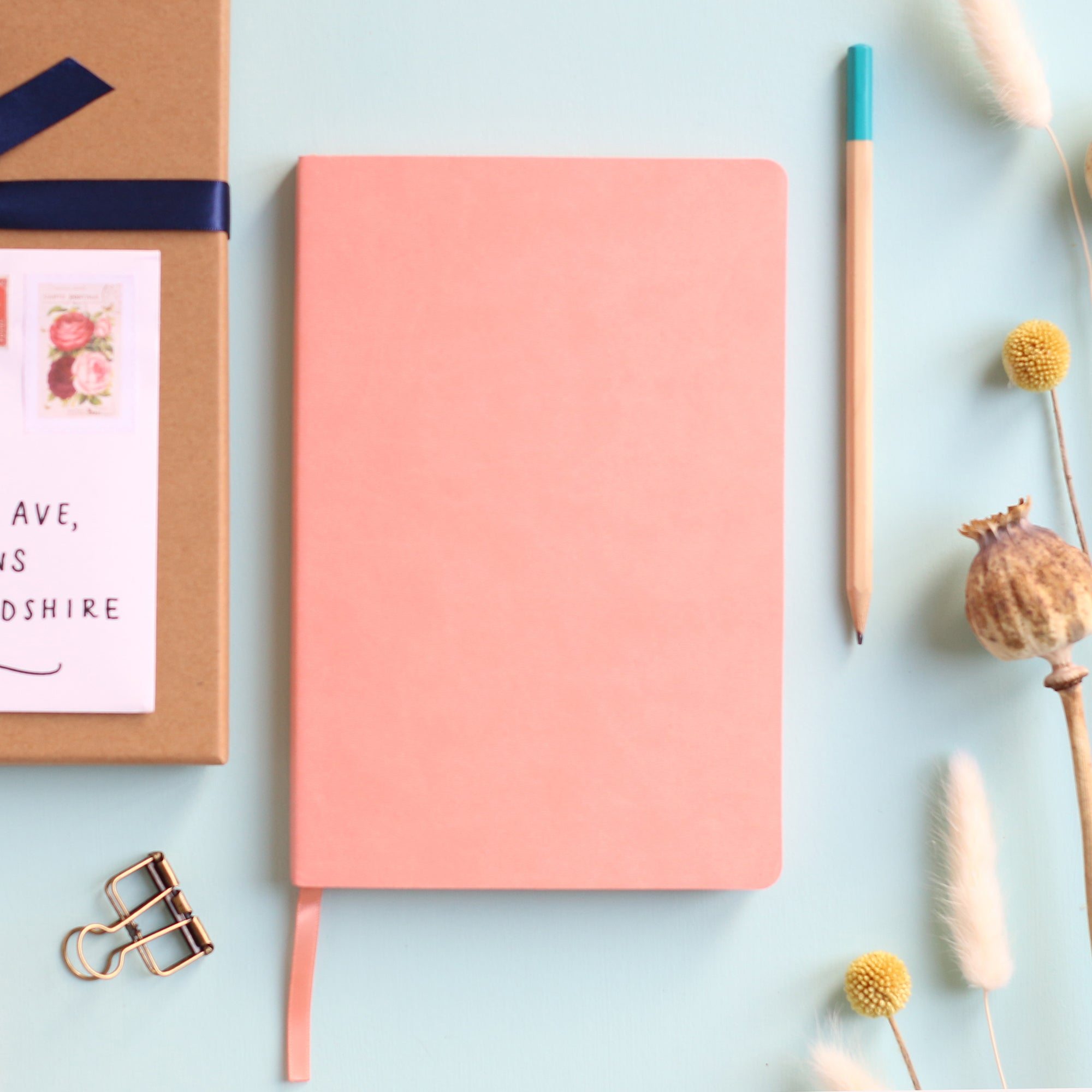 A top down image of a plain simple pink vegan leather notebook. It is resting on a pale aqua table. Surrounding the book are various stationary items and dried flowers.