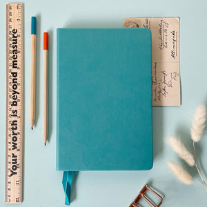 A top down image of a plain simple turquoise vegan leather notebook. It is resting on a light blue surface. Surrounding the book are various stationary items and dried flowers.