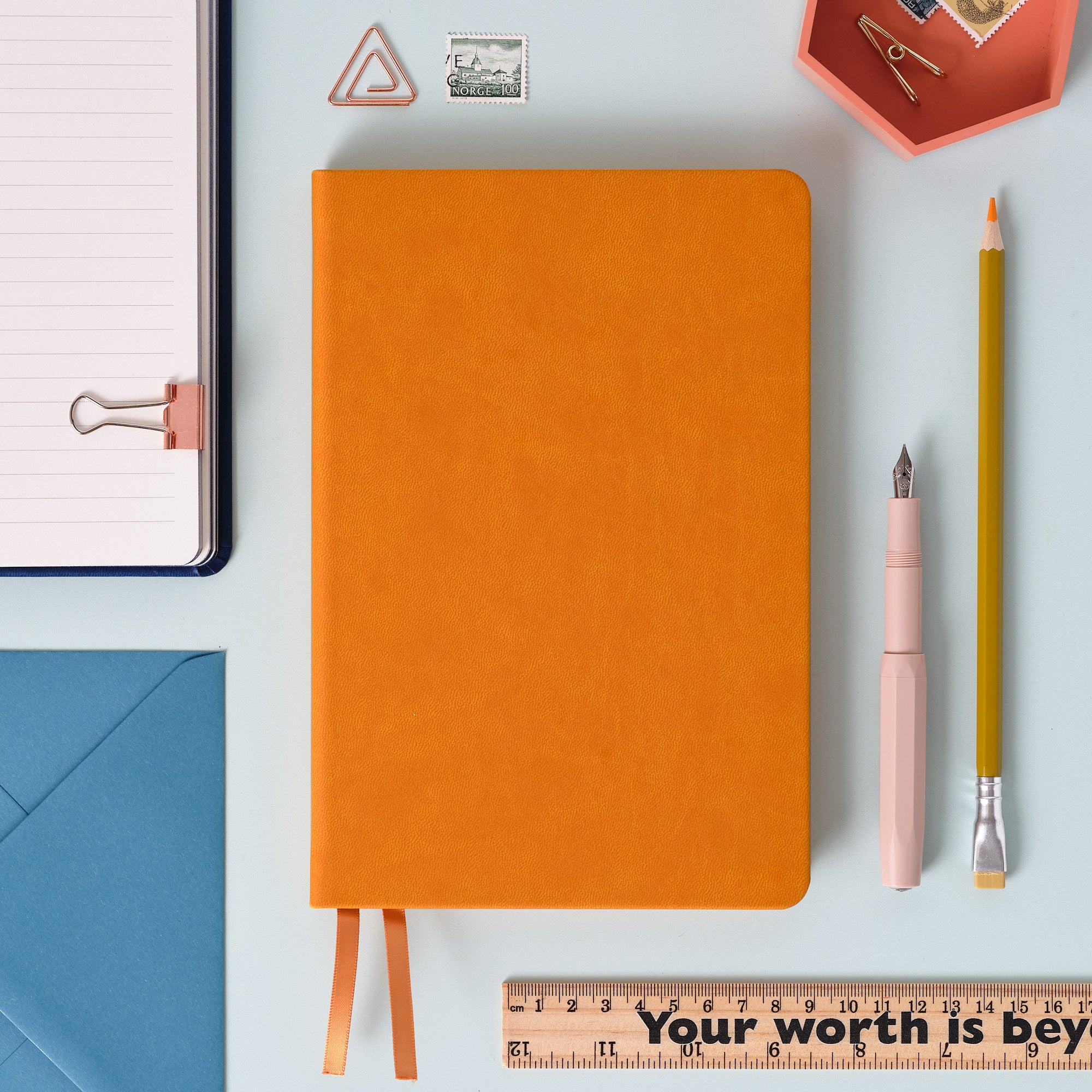 A top down image of a plain simple mustard yellow vegan leather notebook. It is resting on a pale aqua table. Surrounding the book are various stationary items.