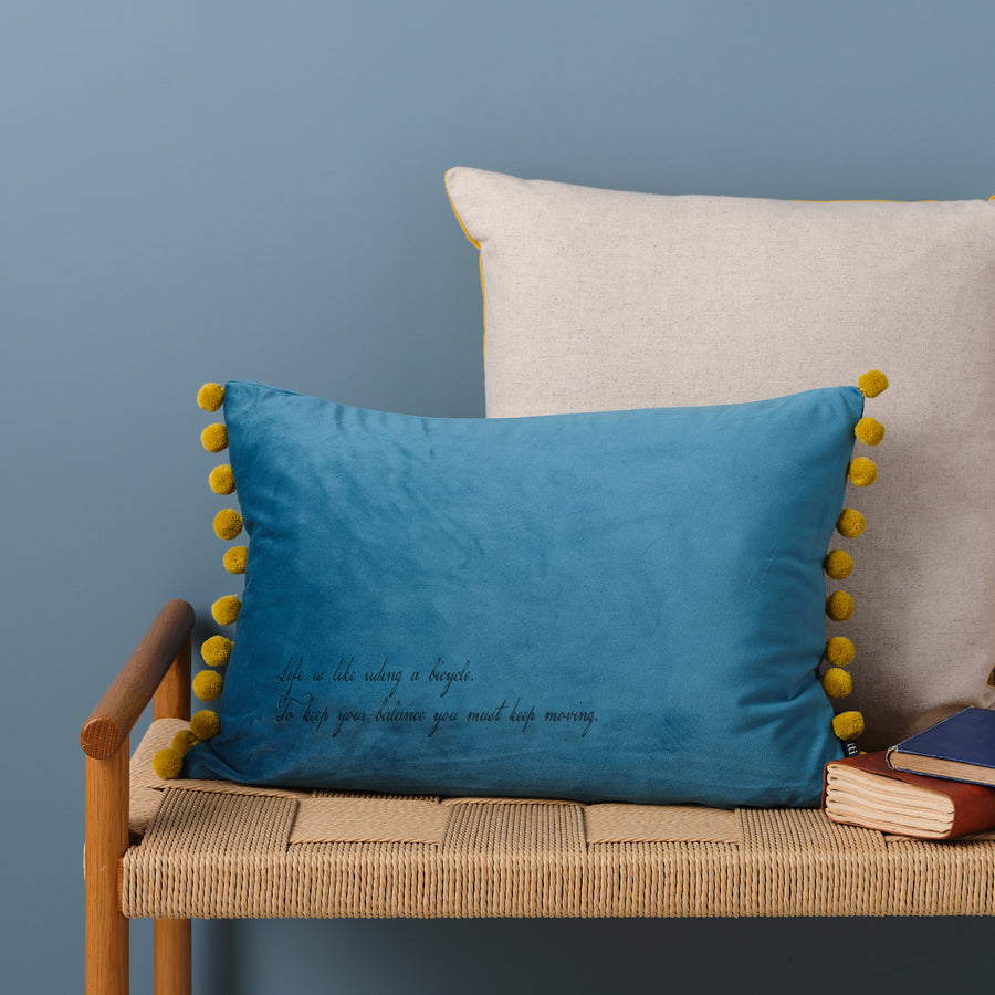 A blue velvet cushion with yellow pom-poms and the quote "Life is like riding a bicycle. To keep your balance you must keep moving" embroidered on it. The cushion is resting on a woven bench with books nearby.