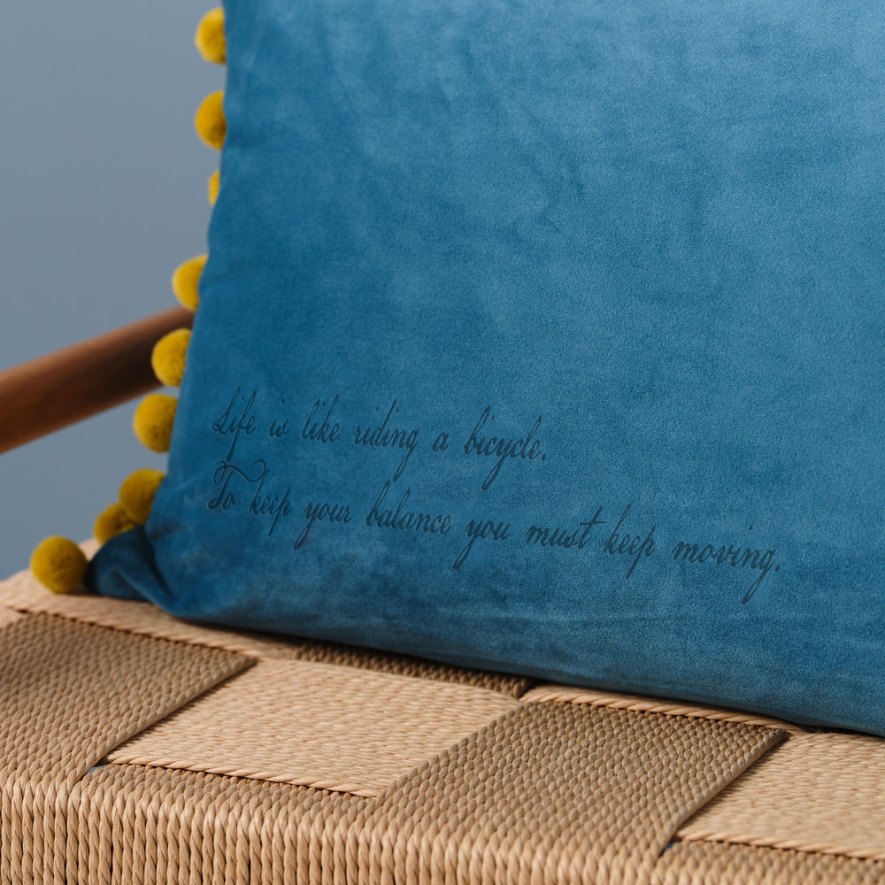 A close-up of a blue velvet cushion with yellow pom-poms. The quote "Life is like riding a bicycle. To keep your balance you must keep moving" is embroidered on the cushion.