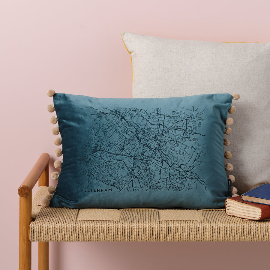  A blue velvet cushion with a map of Cheltenham embroidered on it. The cushion has pom-poms along the edge and is resting on a woven bench with books nearby.