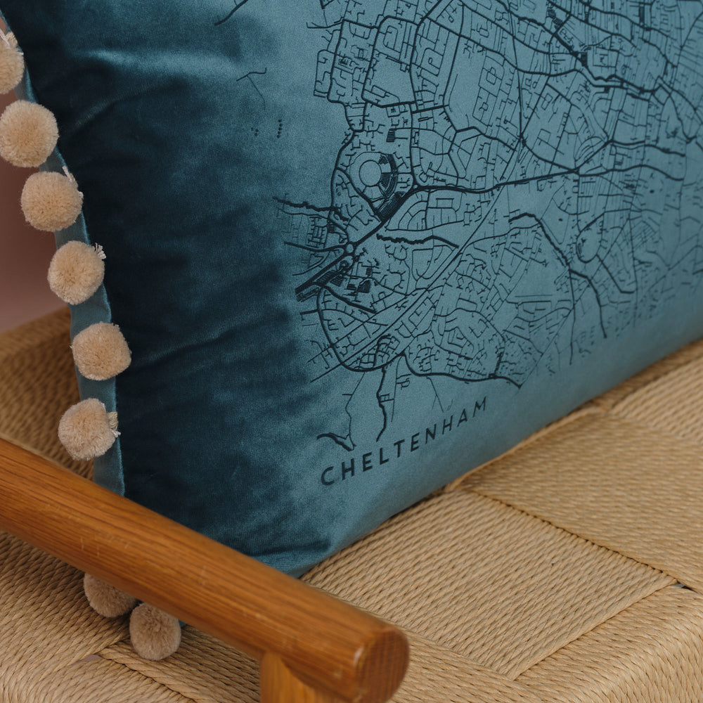 A close-up of a blue velvet cushion with a detailed map of Cheltenham embroidered on it. The cushion has beige pom-poms along the edge and is resting on a wooden chair with a woven seat.