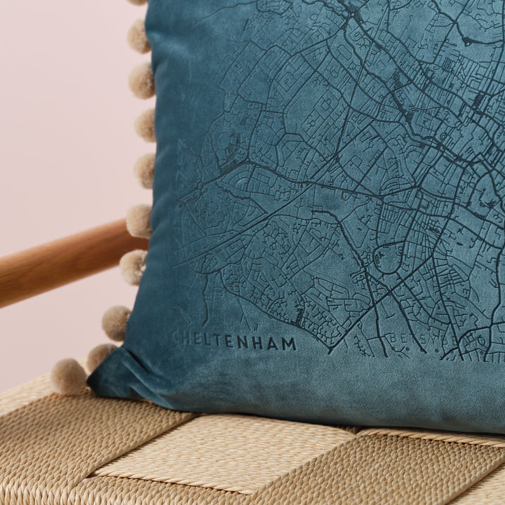 A close-up of a blue velvet cushion with a detailed map of Cheltenham embroidered on it. The cushion has beige pom-poms along the edge and is resting on a wooden chair with a woven seat. The map details are visible, including street names and landmarks.