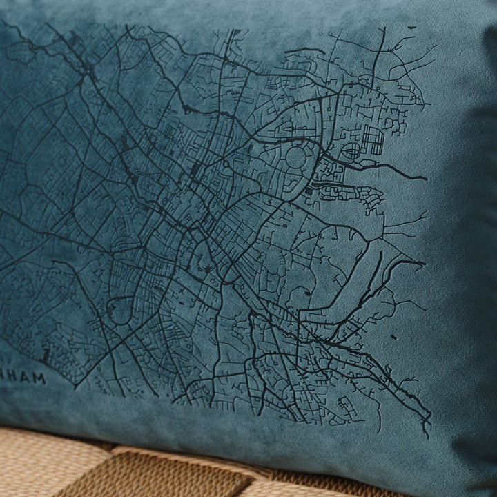 A close-up of a blue velvet cushion with a detailed map of Cheltenham embroidered on it. The cushion has beige pom-poms along the edge and is resting on a wooden chair with a woven seat. The map details are visible, including street names and landmarks.