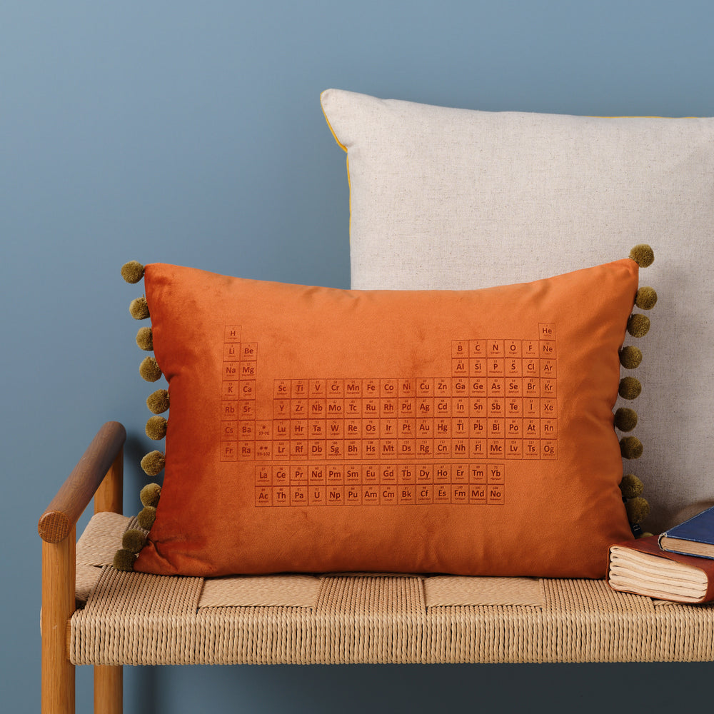 An orange velvet cushion with pom-poms. The periodic table is embroidered on the cushion. It's resting on a woven bench with books nearby.