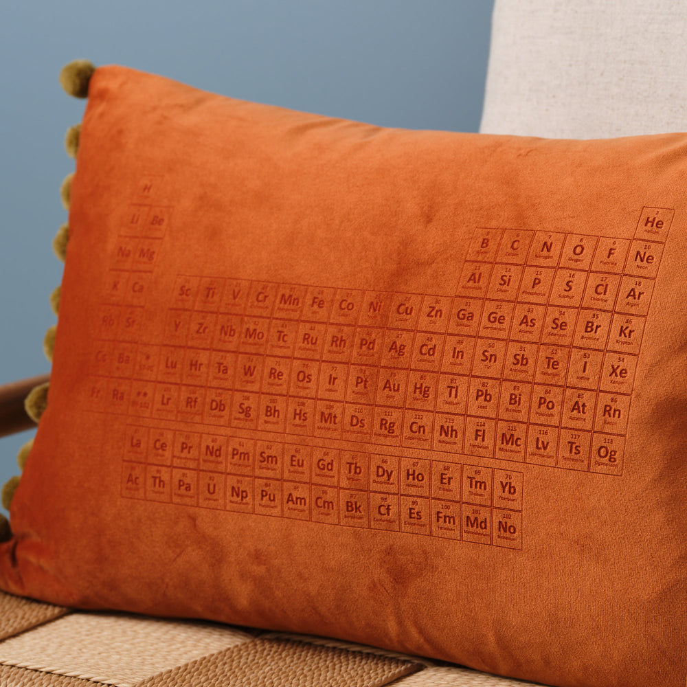 A close-up of an orange velvet cushion with pom-poms. The periodic table is embroidered on the cushion.