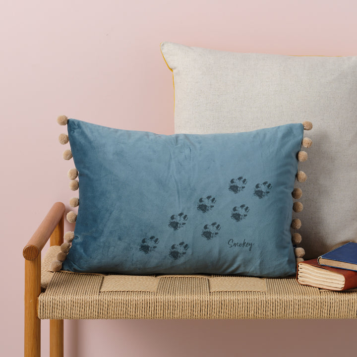 A ocean velvet cushion with bamboo pompoms. There are paw prints and the name Smokey laser engraved on it. The cushion is resting on a woven bench with books nearby.