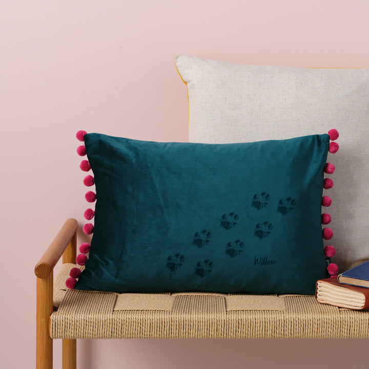 A teal velvet cushion with berry pompoms. There are paw prints and the name Willow laser engraved on it. The cushion is resting on a woven bench with books nearby.
