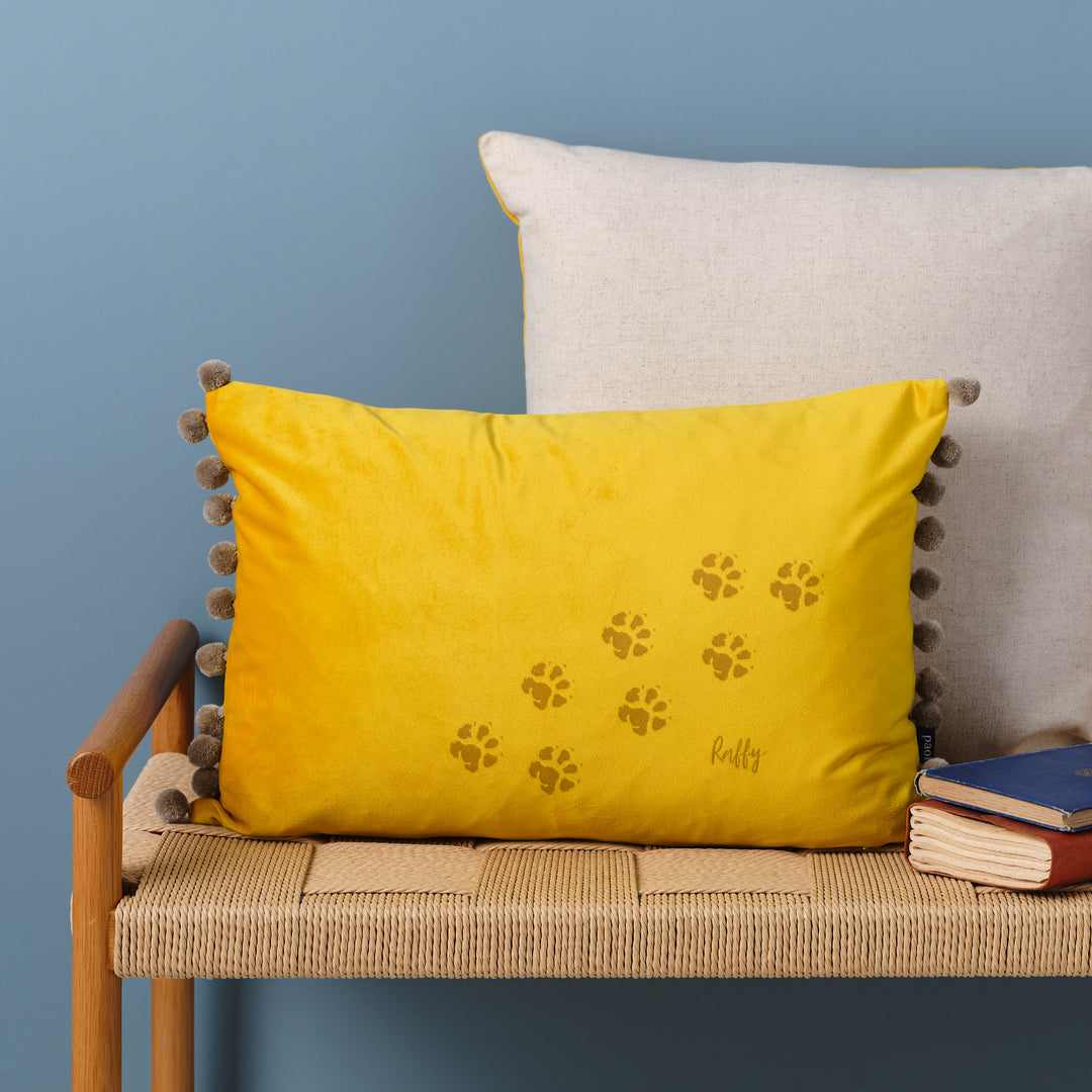 A yellow velvet cushion with bamboo pompoms. There are paw prints and the name Raffy laser engraved on it. The cushion is resting on a woven bench with books nearby.