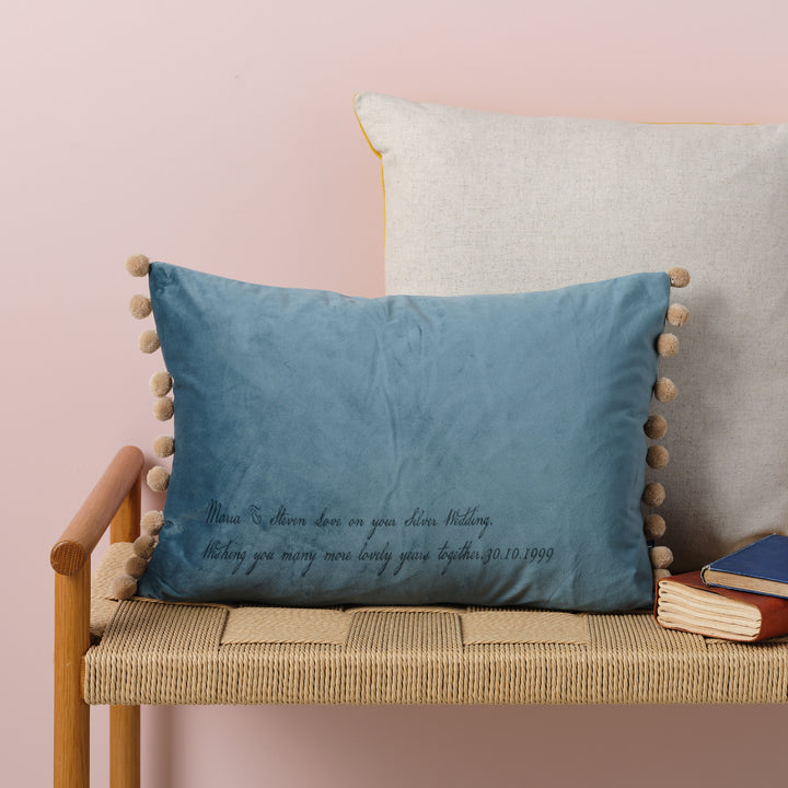 A ocean velvet cushion with bamboo coloured pom-poms and the a quote  laser engraved on it. The cushion is resting on a woven bench with books nearby.