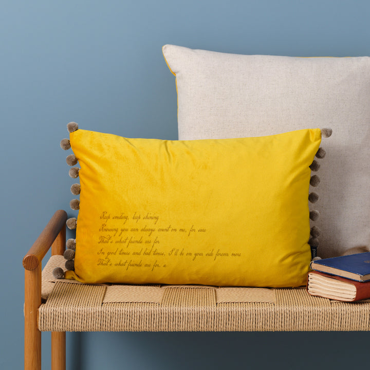 A yellow velvet cushion with bamboo coloured pom-poms and the a quote  laser engraved on it. The cushion is resting on a woven bench with books nearby.
