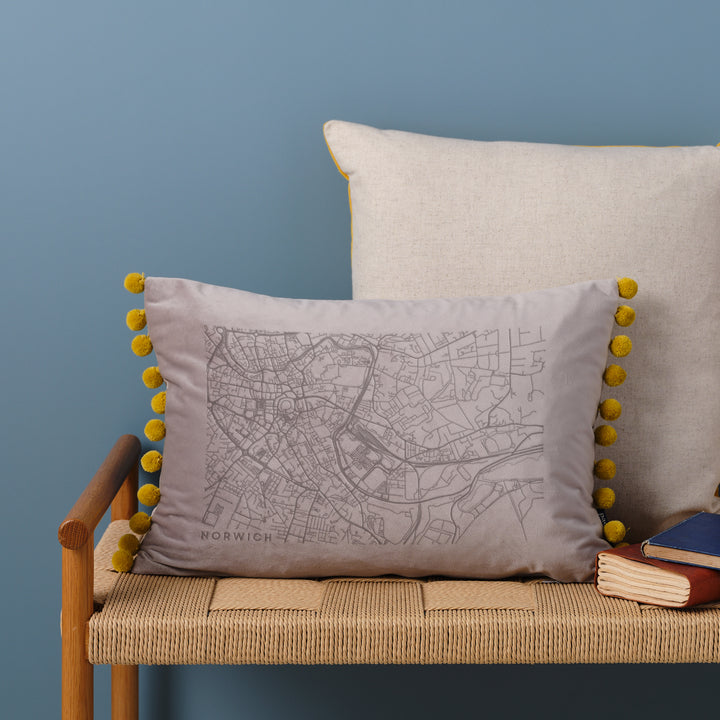  A silver velvet cushion with a map engraved on it. The cushion has pom-poms along the edge and is resting on a woven bench with books nearby.