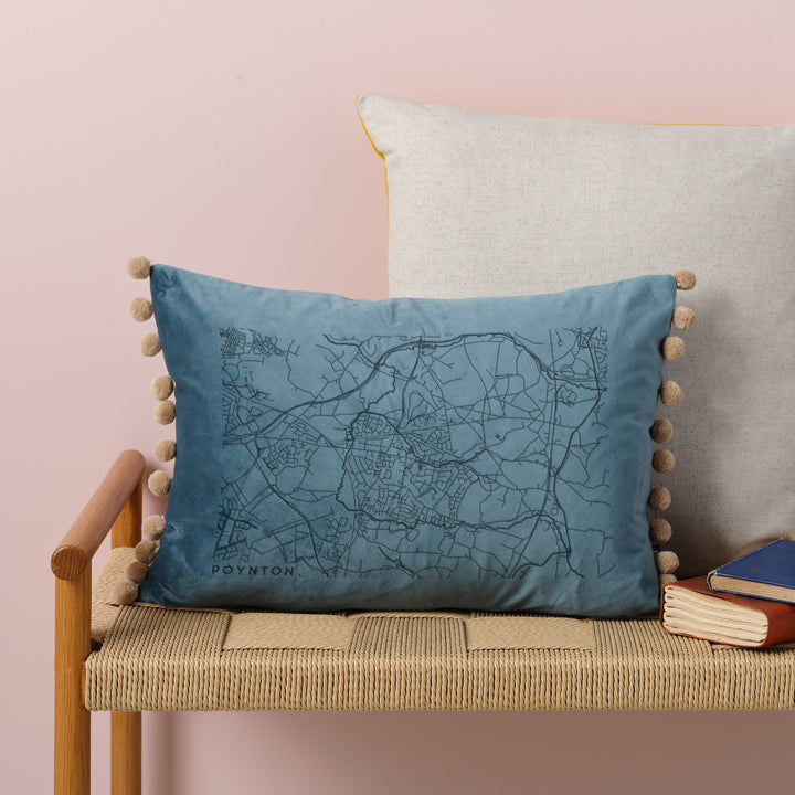  A ovean velvet cushion with a map engraved on it. The cushion has pom-poms along the edge and is resting on a woven bench with books nearby.