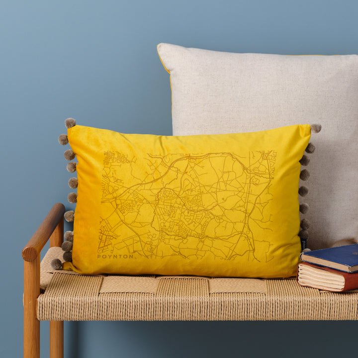  A yellow velvet cushion with a map engraved on it. The cushion has pom-poms along the edge and is resting on a woven bench with books nearby.