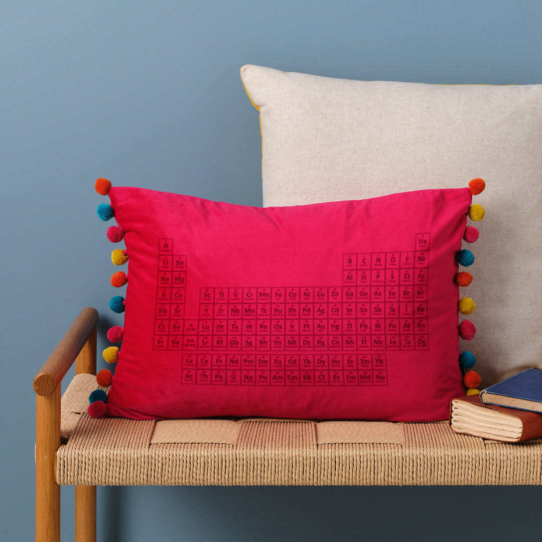 An hot pink velvet cushion with pom-poms. The periodic table is embroidered on the cushion. It's resting on a woven bench with books nearby.