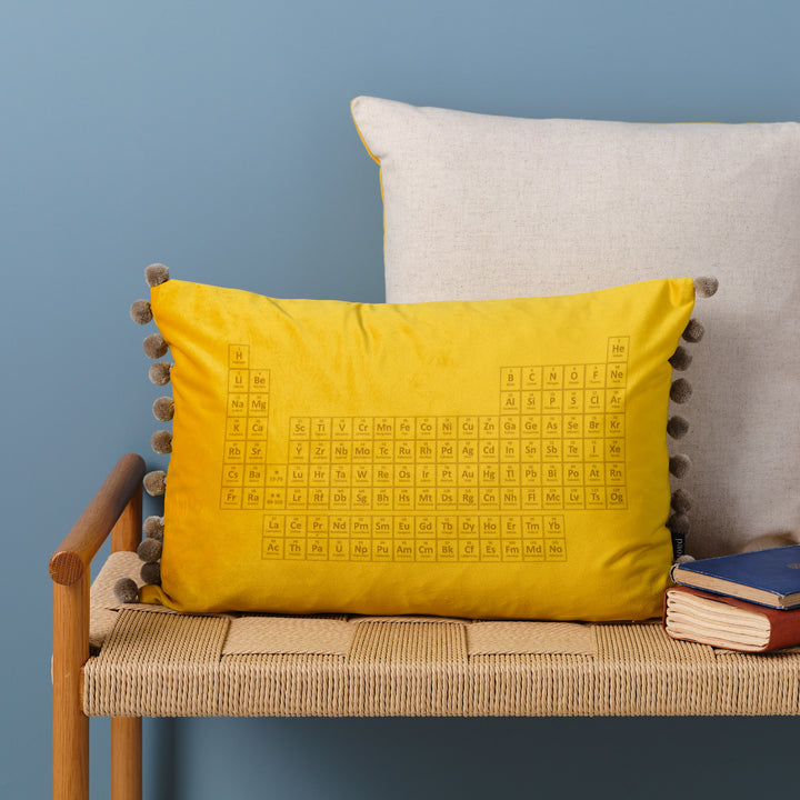 An yellow velvet cushion with pom-poms. The periodic table is embroidered on the cushion. It's resting on a woven bench with books nearby.