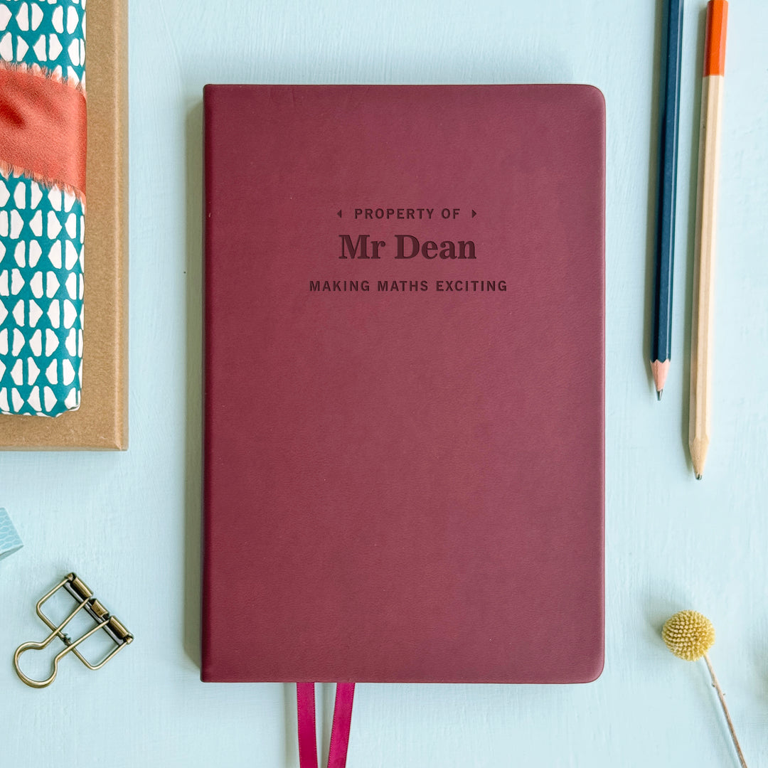 Maroon notebook with 'Property of Mr Dean Making Maths Exciting' text on a light blue background with stationery items.