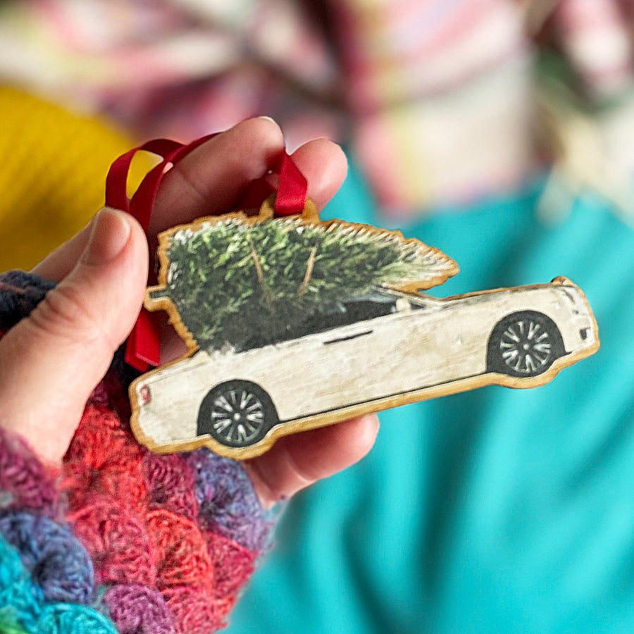 A hand holding a wooden Christmas tree decoration featuring a Rolls Royce car with a Christmas tree on the roof.