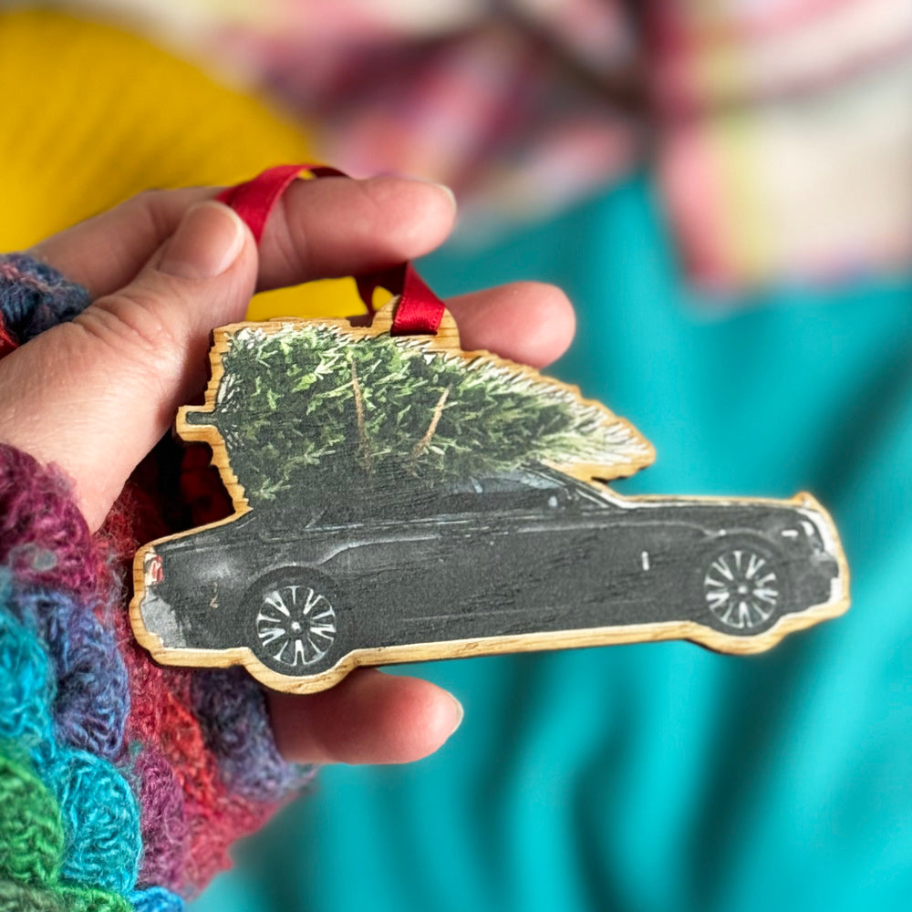 A hand holding a wooden Christmas tree decoration gift featuring a black Rolls Royce car with a Christmas tree on the roof.