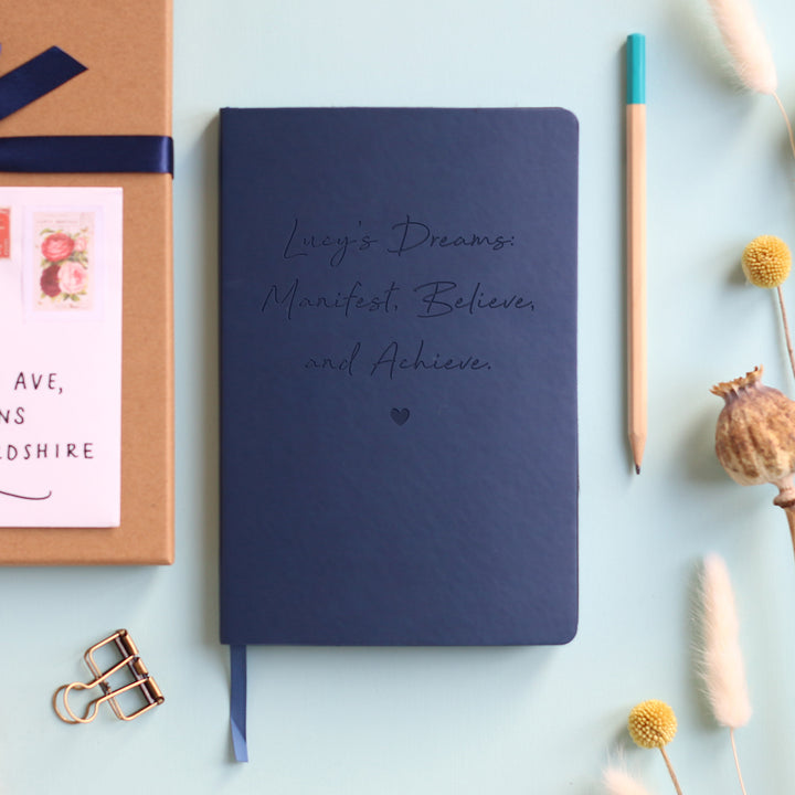 A top down image of a navy blue vegan leather notebook resting on a pale aqua table. The front of the notebook is engraved with a heart and the phrase "Lucy’s Dreams. Manifest, Believe and Achieve" in a script font. Surrounding the book are various stationary items and dried flowers.