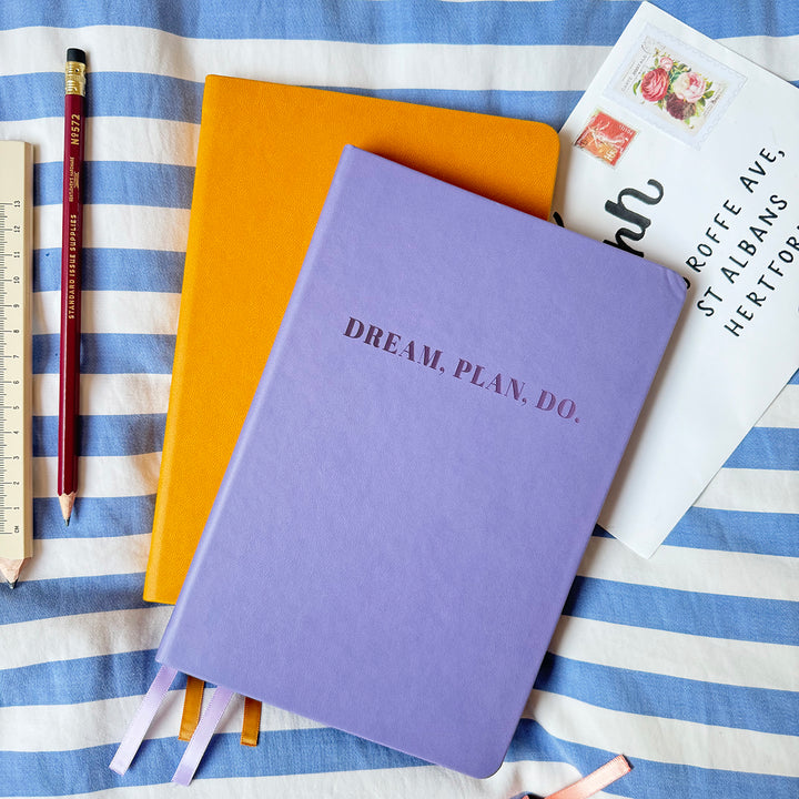 A top down image of two vegan leather notebooks, a lilac notebook on top of a yellow book. They are resting on a blue and white striped tablecloth. The lilac journal is personalised with the text Dream, Plan, Do. Surrounding the book are various stationary items.