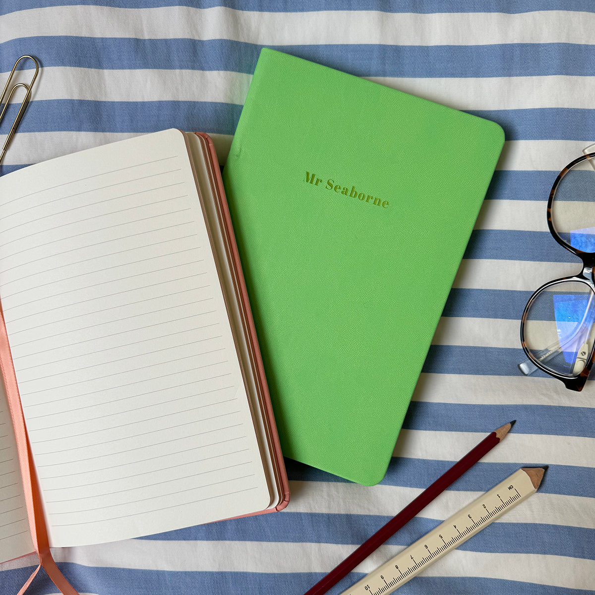 A top down image of two vegan leather notebooks, a green notebook and an open pink journal. They are resting on a blue and white striped tablecloth. The green notebook journal is personalised with the engraving Mr Seaborne. Surrounding the books are various stationary items.