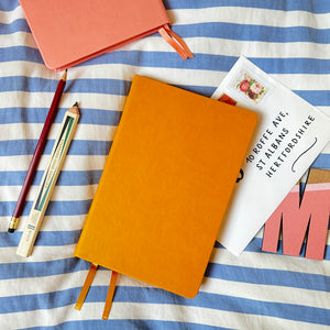 Two blank notebooks, one mustard yellow and one pink, lie on a striped blue and white fabric. A pencil, a postcard with an address, and a wooden letter "M" are scattered around. The postcard has a floral stamp.