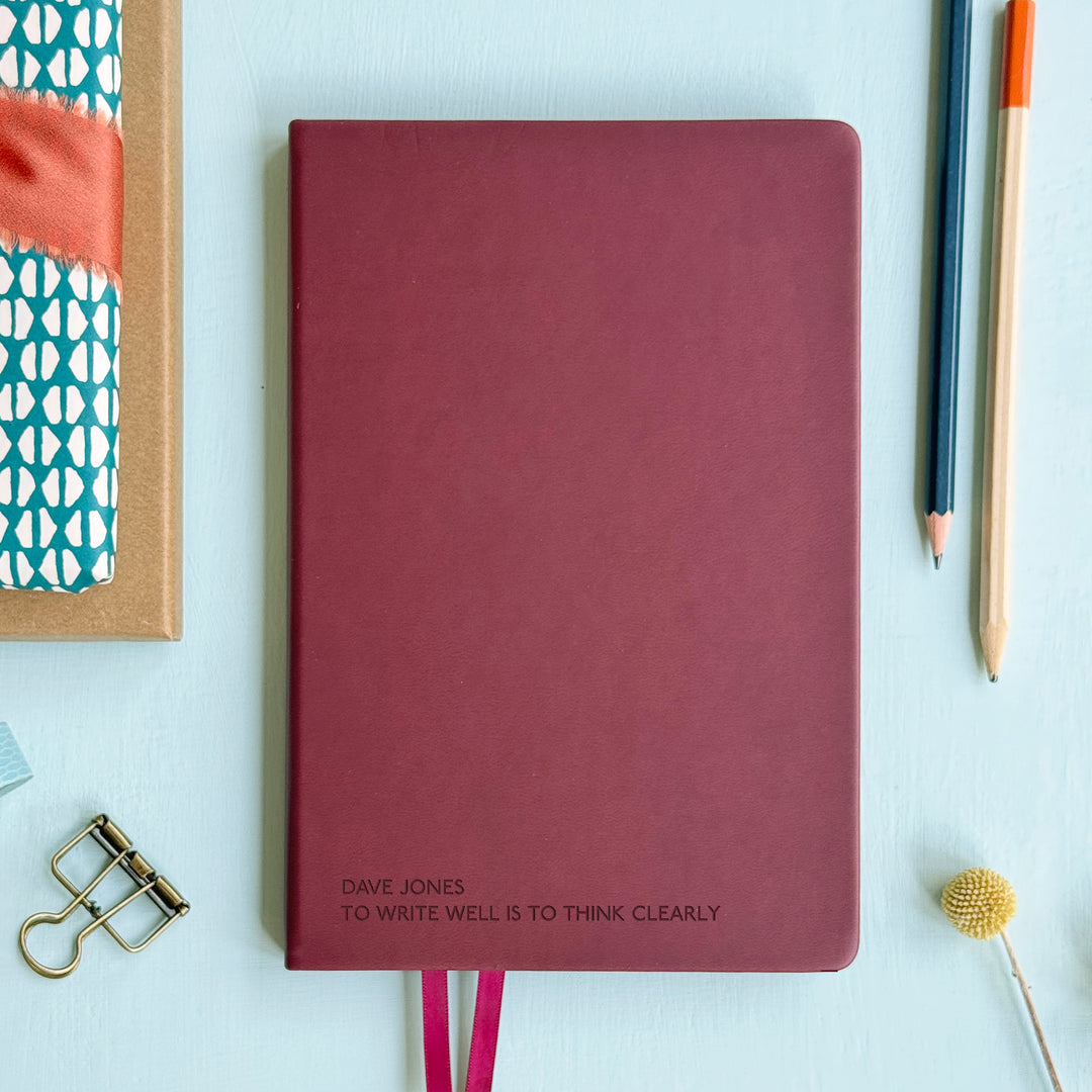 A top down image of a burgundy hardcover vegan leather notebook resting on a pale aqua table. The bottom of the book is engraved with the personalised message  - Dave Jones - To write well is to think clearly. Surrounding the book are various stationary items and dried flowers.