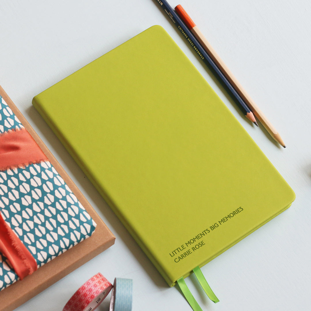 A top down image of a lime green hardcover vegan leather notebook resting on a pale aqua table. The bottom of the book is engraved with the personalised message ' Little moments big memories Carrie Rose' Surrounding the book are various stationary items.