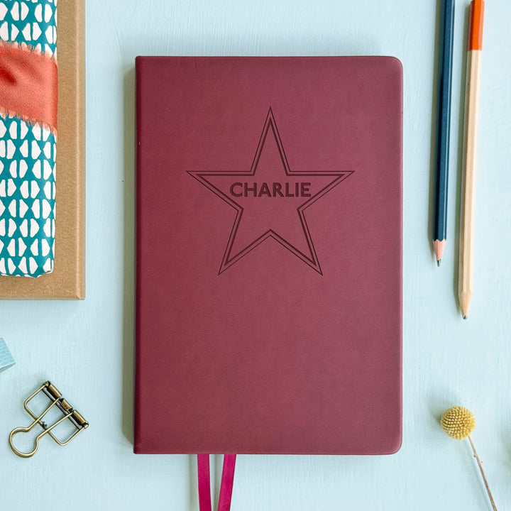 A flat lay image of a burgundy vegan leather notebook resting on a pale aqua table. The front of the notebook is engraved with a large 5 point star and is personalised with the name Charlie. Surrounding the book are various stationary items and dried flowers.
