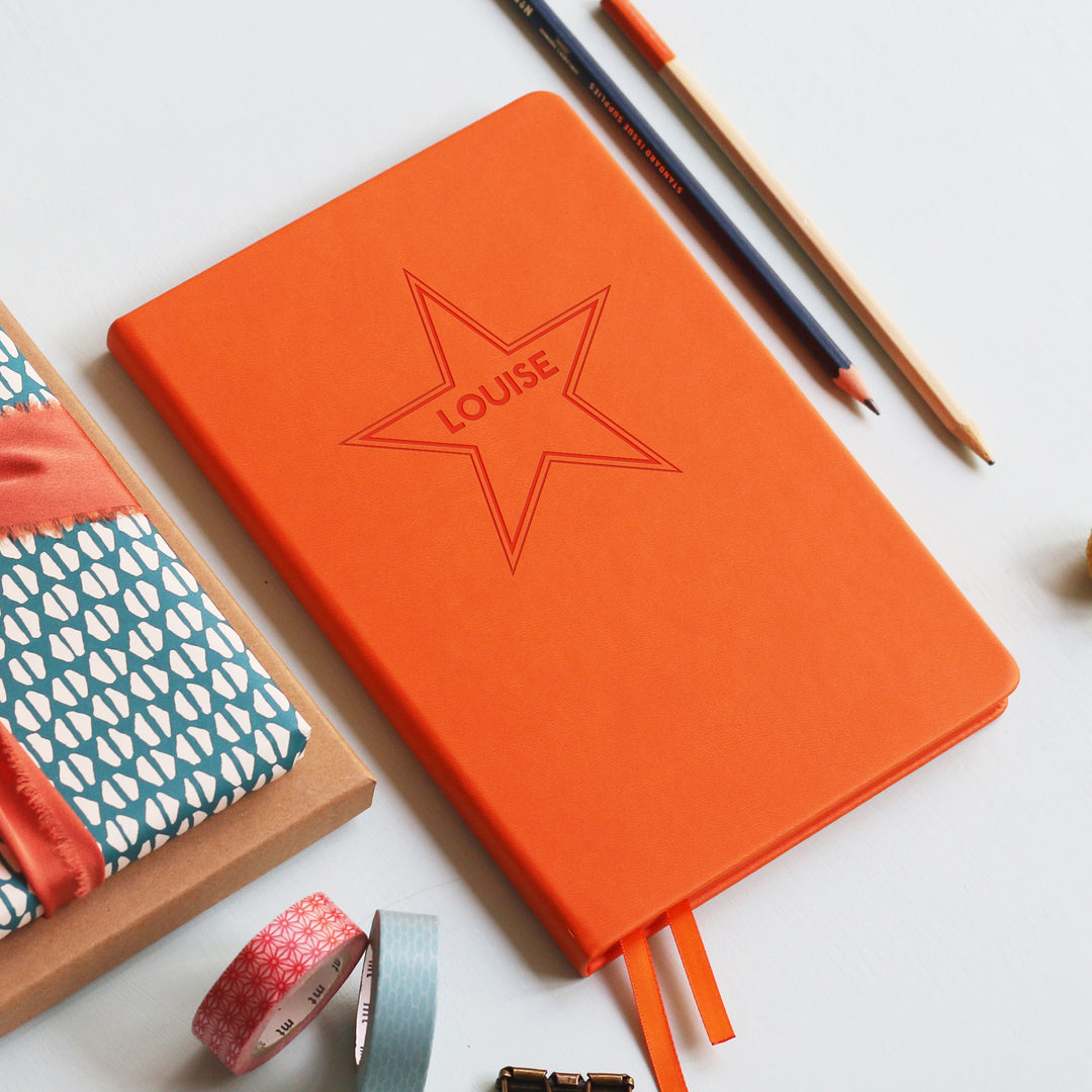 A flat lay image of a orange vegan leather notebook resting on a pale aqua table. The front of the notebook is engraved with a large 5 point star and is personalised with the name Louise. Surrounding the book are various stationary items.
