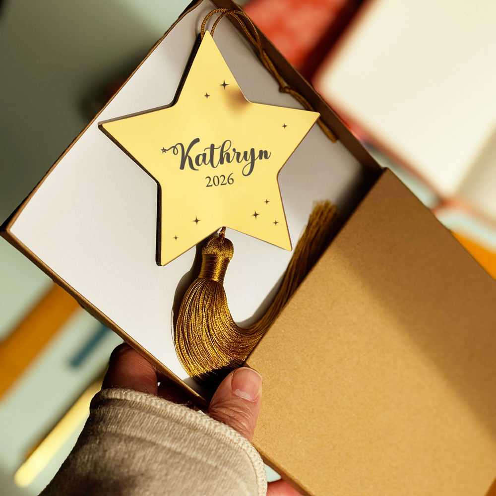 Metallic Gold star-shaped baby Christmas tree decoration with engraved names and tassel held by a hand in a brown presentation gift box.