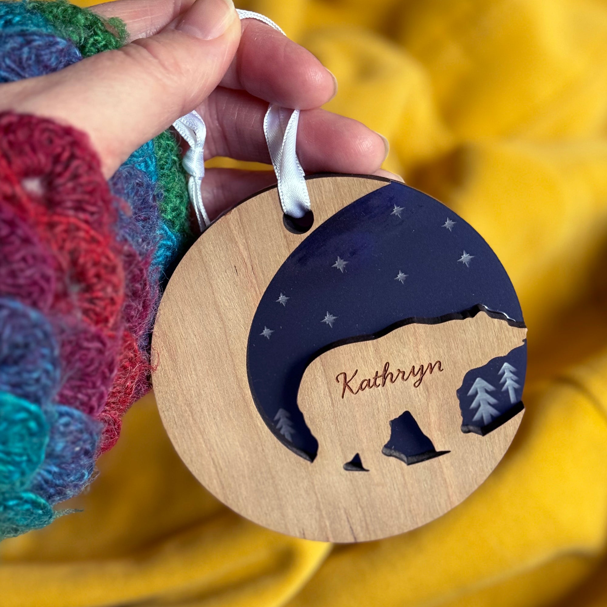 A hand holds a wooden Christmas ornament with a bear silhouette and the name "Kathryn" engraved. The ornament has a crescent moon cutout and a dark blue inset with stars. The hand is wearing a colorful knitted glove.