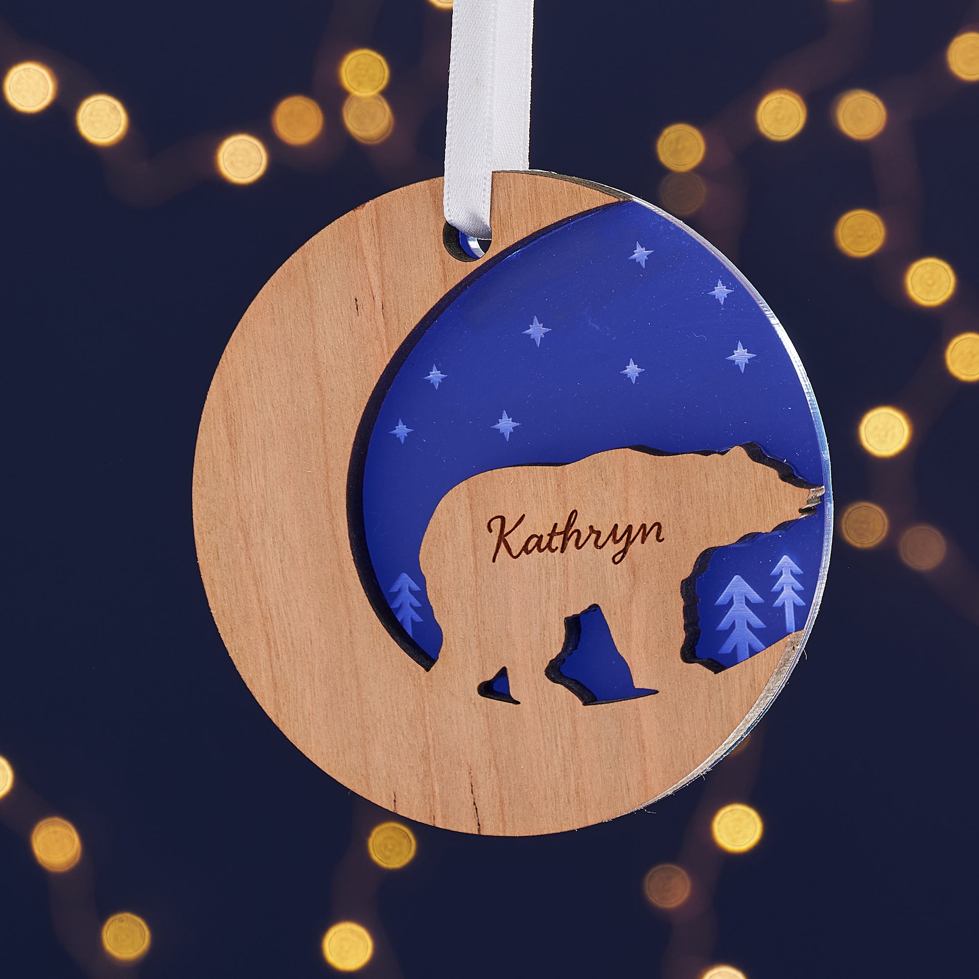  Two wooden Christmas ornaments, one a crescent moon and the other a circle, hang from a branch against a dark blue background with bokeh lights. Each ornament features a cutout silhouette of a bear and is engraved with a name, "Elizabeth" and "Kathryn."