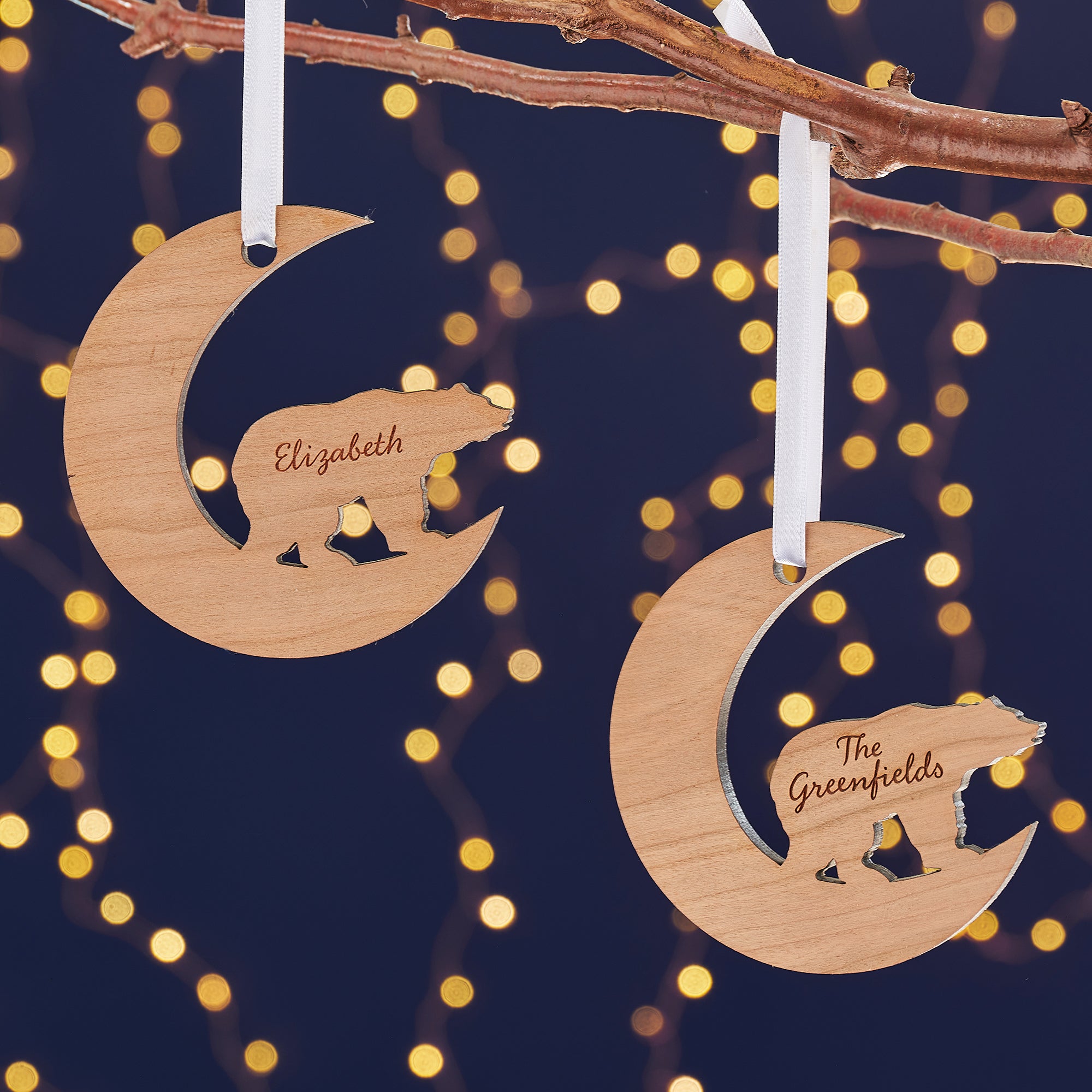 Two crescent moon-shaped wooden Christmas ornaments hang from a branch against a dark blue background with bokeh lights. Each ornament features a cutout silhouette of a bear and is engraved with a name, "Elizabeth" and "The Greenfields."