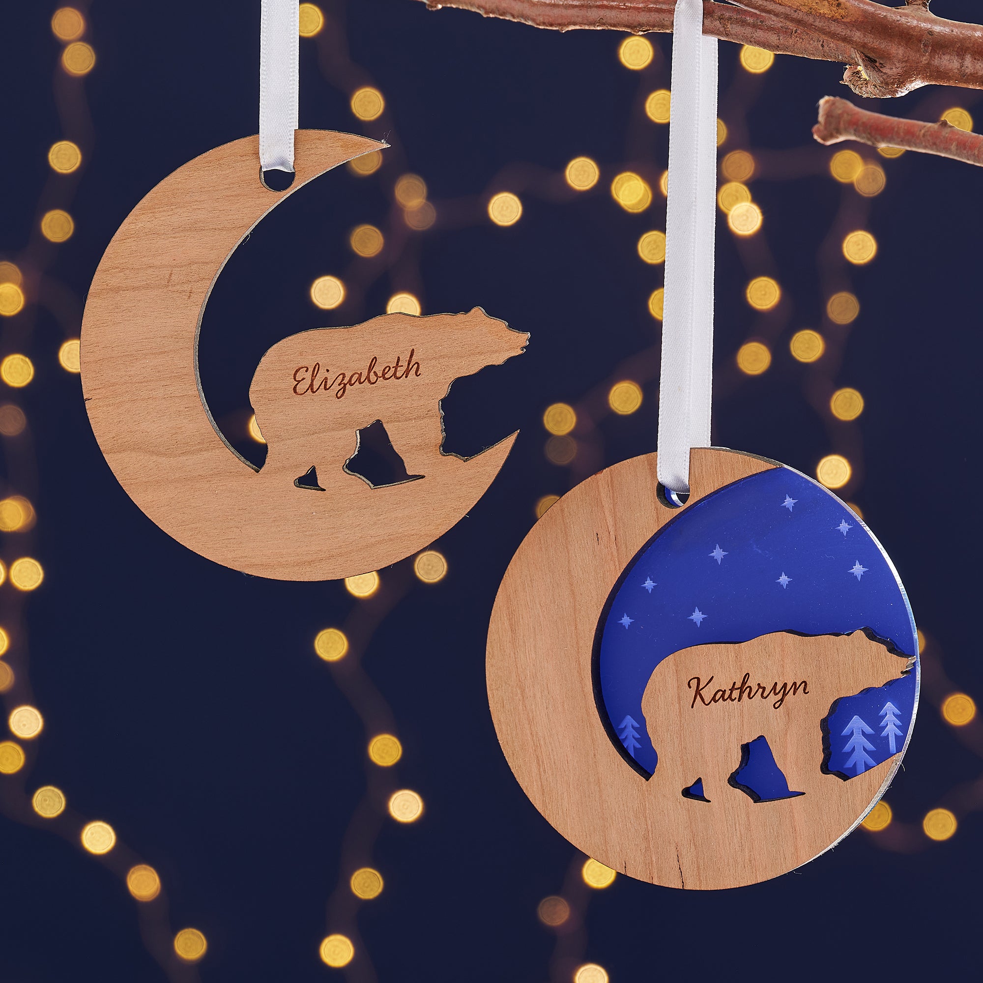  Two wooden Christmas ornaments, one a crescent moon and the other a circle, hang from a branch against a dark blue background with bokeh lights. Each ornament features a cutout silhouette of a bear and is engraved with a name, "Elizabeth" and "Kathryn."