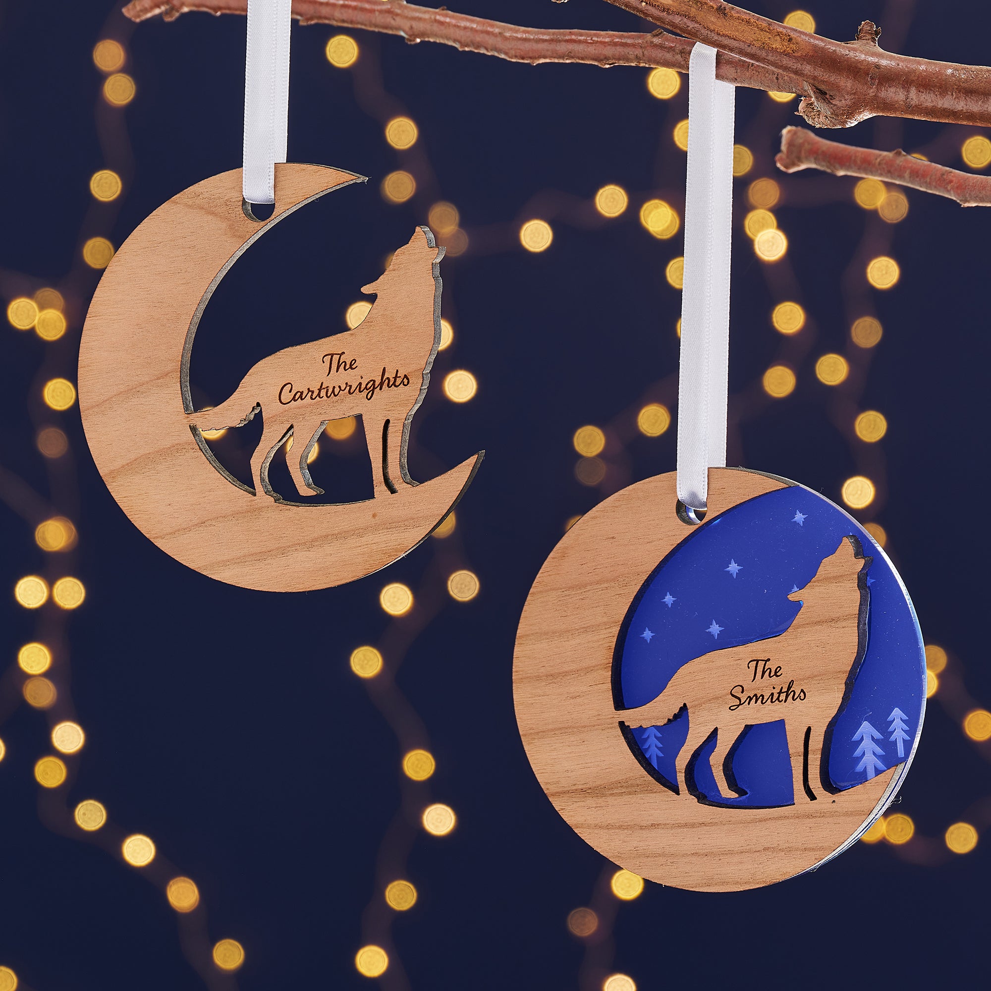 Two wooden Christmas ornaments, one a crescent moon and the other a circle, hang from a branch against a dark blue background with bokeh lights. Each ornament features a cutout silhouette of a wolf howling at the moon and is engraved with a family name, "The Cartwrights" and "The Smiths."